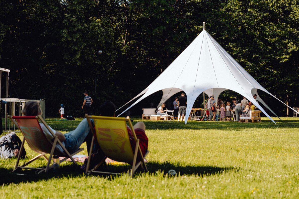 A large white tent is set up on a grassy field, where people have gathered. In the foreground, two people are lounging on deck chairs opposite the tent. The area is surrounded by trees, and the sun shines brightly, creating a lively and casual atmosphere - the perfect scene for a photo essay of the event.  