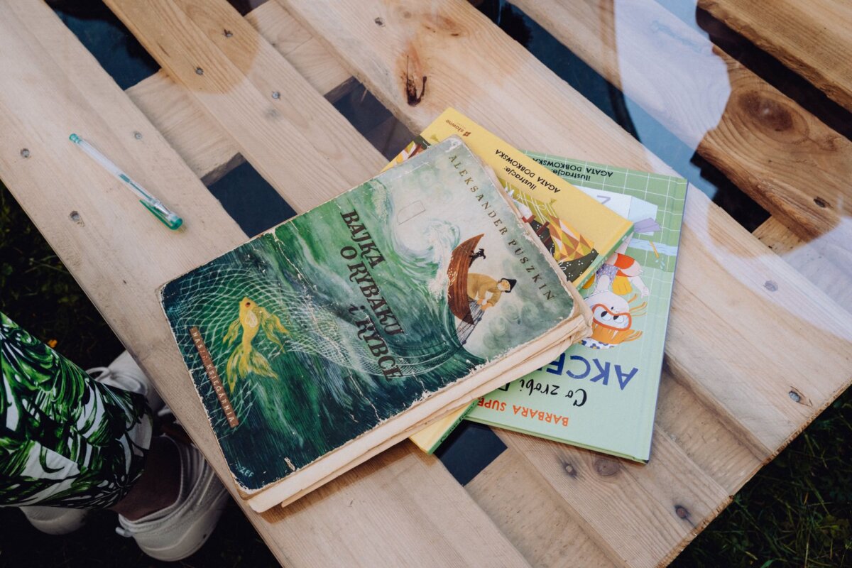 Taken by a photographer from Warsaw, a close-up of several children's books on a wooden surface. The upper book features an illustration of a person catching a fish in a net from a boat, with text written in Cyrillic. Also visible is part of a foot in a tennis shoe and a toothbrush.  