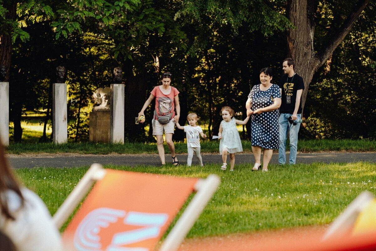 A group of three adults and two children are walking in a park with lush green grass and tall trees. Two adults are holding hands with a little girl in a white dress, while a third adult is leading another child. The picturesque moment, perfect for any event photographer warsaw, also features two orange deck chairs in the foreground.  