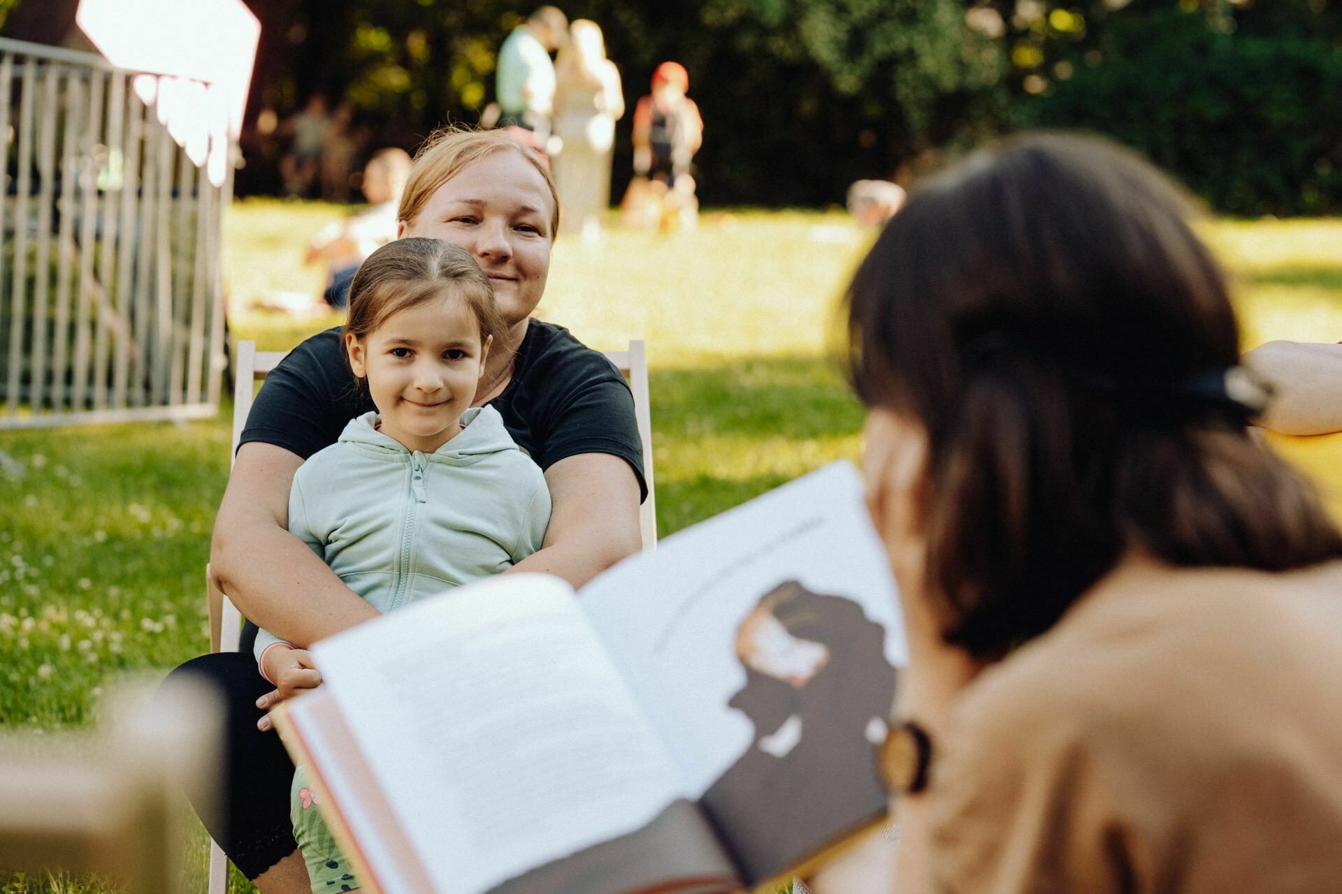 A woman sits outside with a young girl on her lap and smiles at someone reading a book in the foreground. The scene takes place in a park, with a backdrop of people and greenery, which was beautifully captured by event photographer Warsaw. 