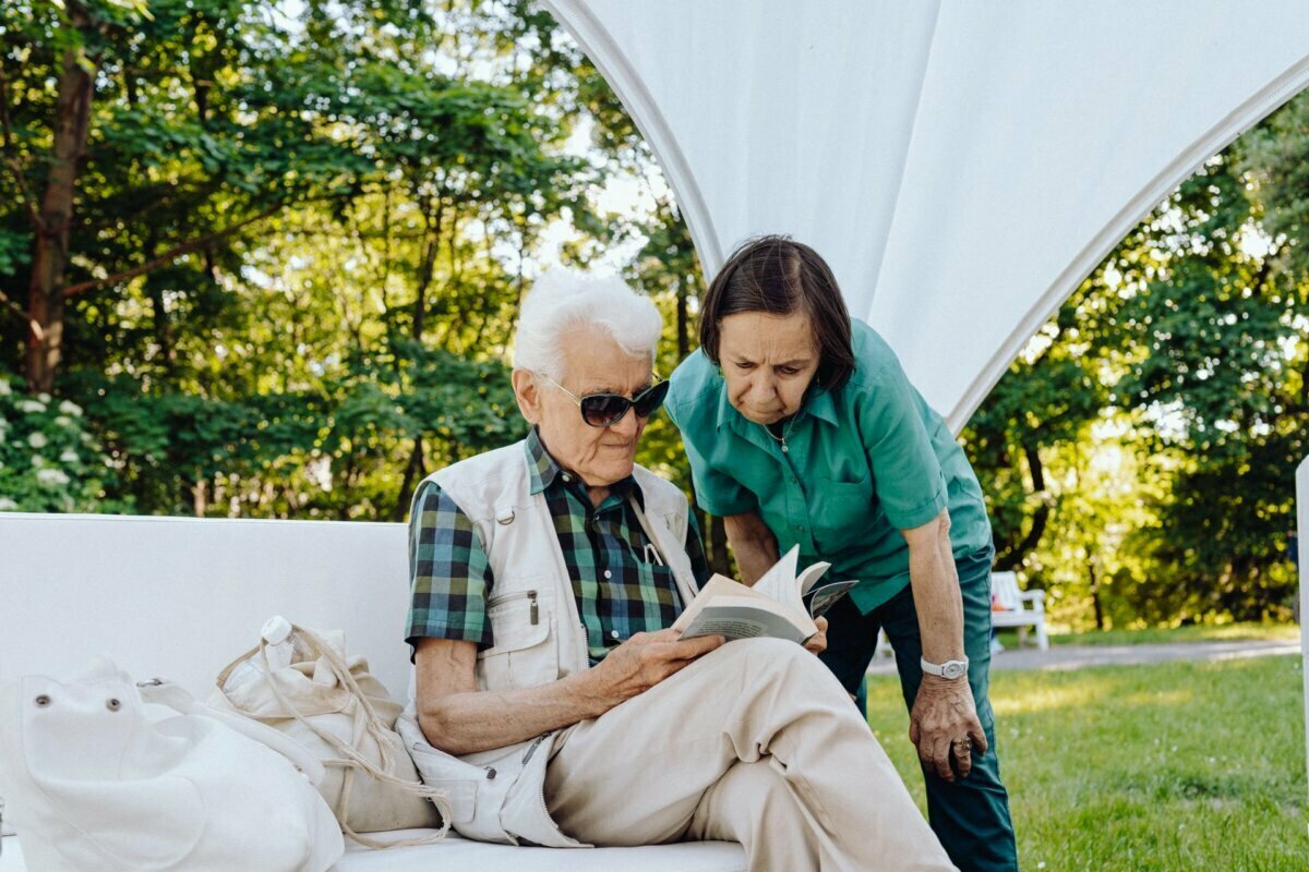 An older man with white hair, wearing sunglasses, is reading a book while sitting on a white bench outside. A woman with short brown hair wearing a green shirt leans over to look at the book. They are in a lush, sunny park with trees in the background - the perfect scene for any Warsaw photographer capturing everyday moments.  