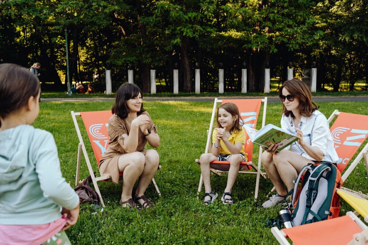 A group of people are sitting on deck chairs in a park. The adult on the right is reading a book to a child. Nearby is another adult and a child engaged in conversation. In the foreground, the child is standing with his back to the camera. The scene is surrounded by trees and greenery, beautifully captured by a photographer from Warsaw.    