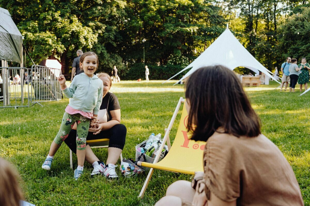 A cheerful young girl in colorful clothes stands near a seated woman on a grassy lawn in a park. The woman, partially obscured, is sitting on a yellow deck chair. A white tent and other people can be seen in the background between the trees. The sunny, vibrant scene captures the essence of the event photo report by the Warsaw-based photographer.   