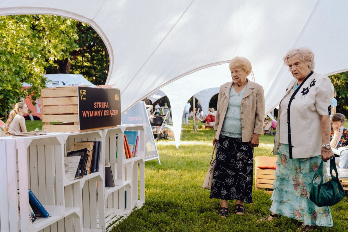 Two elderly women are standing near a book exchange point set up outside under a white canopy. They are looking at books on wooden shelves made of crates. In the background, people are sitting on benches and chairs, enjoying the sunny day of the event, beautifully captured by event photographer warsaw.  