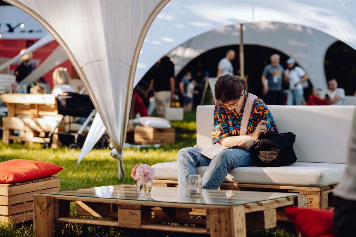 A person sits on a white sofa and reads a book outdoors, beautifully captured by *event photographer Warsaw*. The area is shaded by large tent structures, while people stand and walk around in the background. Wooden pallets serve as tables and seating, creating a casual atmosphere.  