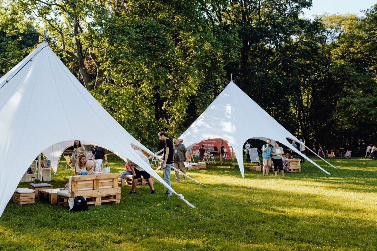 People enjoy a relaxed outdoor event on a grassy area with large triangular white tents. The stage is ideal for photo coverage of the event, equipped with wooden pallet seating and a variety of attractions, surrounded by lush trees and greenery. The atmosphere is casual and inviting.  