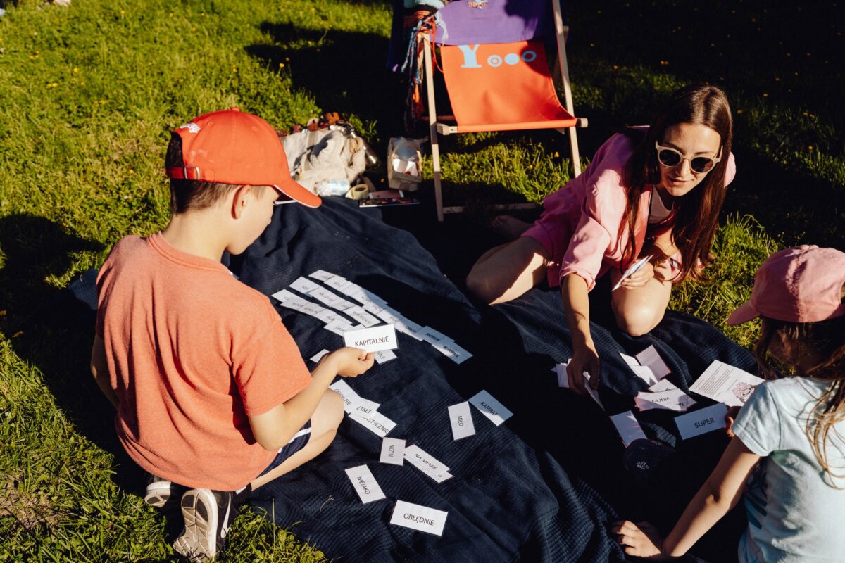 A woman and two children are sitting on a blue blanket on a grassy ground, engrossed in some activity, and various cards are spread out in front of them. The woman wears sunglasses and the children wear baseball caps. In the background is an orange chair with "Yooo" printed on it, which adds a charming touch to the event photo.  