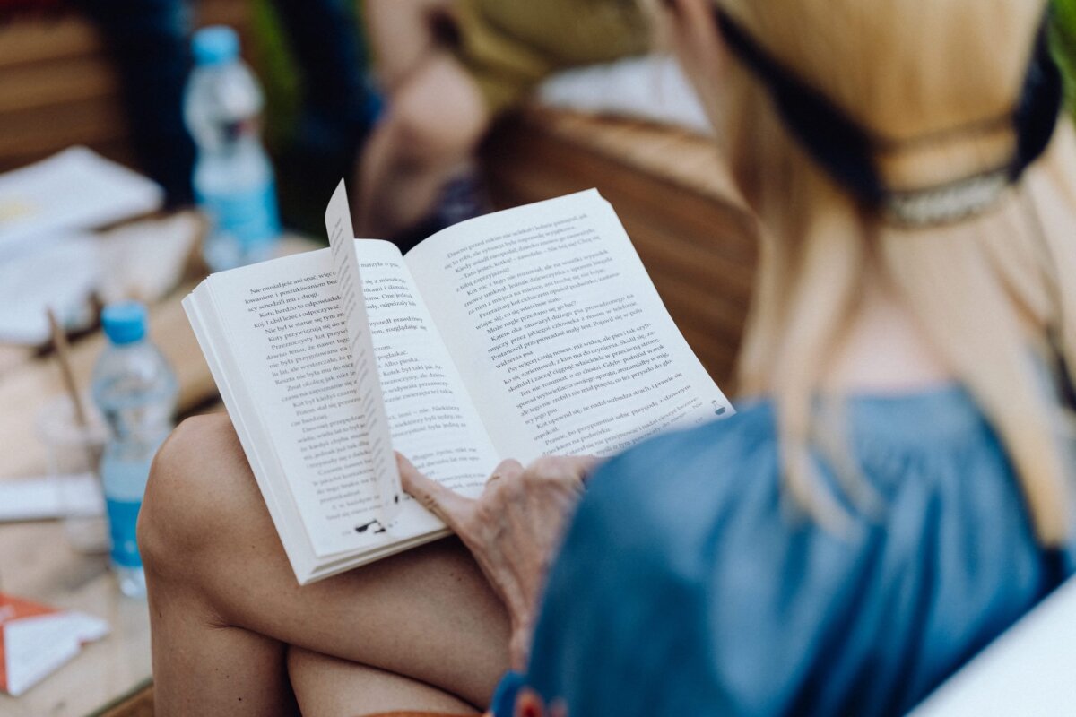 A person with long, blonde hair reads a book while sitting during a photo-op of the event. The text on the open book appears to be written in English. On the table in front of them lies a plastic bottle of water and other items. Dressed in casual blue attire, they appear immersed in reading.   