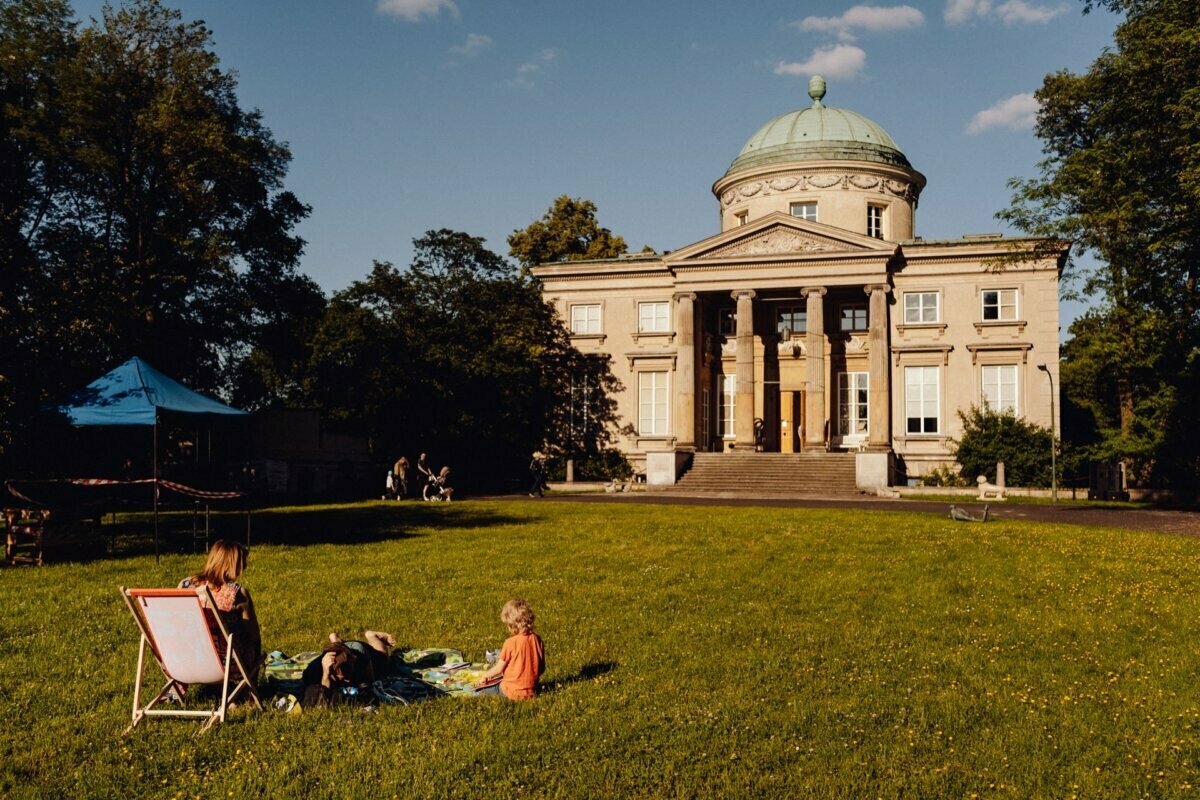 A stately neoclassical mansion with a cupola stands at the edge of a lush lawn bathed in sunlight. In the foreground, a relaxed group of people is enjoying a picnic on a blanket and deck chair, presumably captured by the event photographer. The trees and distant tent structure are visible under a blue sky with a few clouds.  