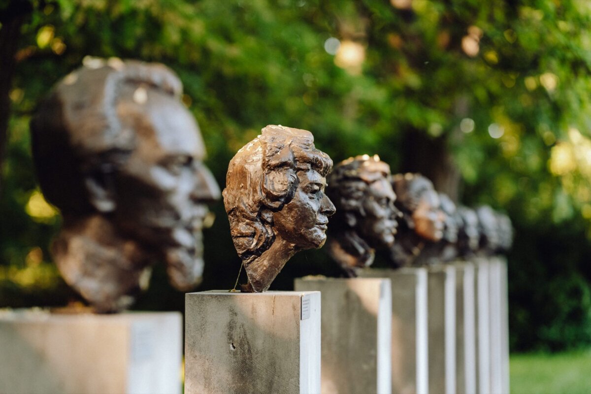 A row of bronze busts is displayed on individual pedestals in the open air. The busts feature different facial expressions and hairstyles. Lush green trees serve as a blurred backdrop, suggesting a location in a park or garden - perfect for a Warsaw-based photographer who can capture stunning visuals.  