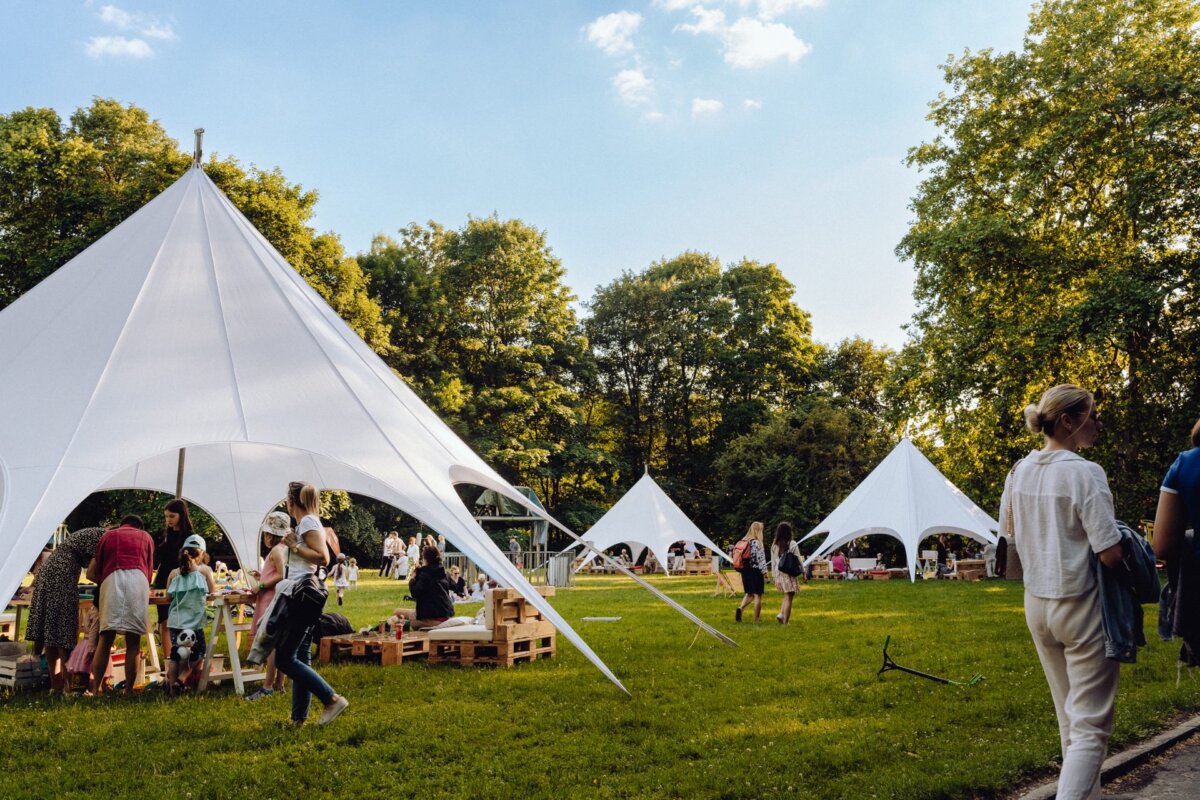 People are enjoying a sunny day at an outdoor event in a park with large white conical tents. Some walk around, others sit on wooden benches under the tents. A photographer from Warsaw captures the lush green grass and surrounding trees, which enhance the festive and casual atmosphere.  