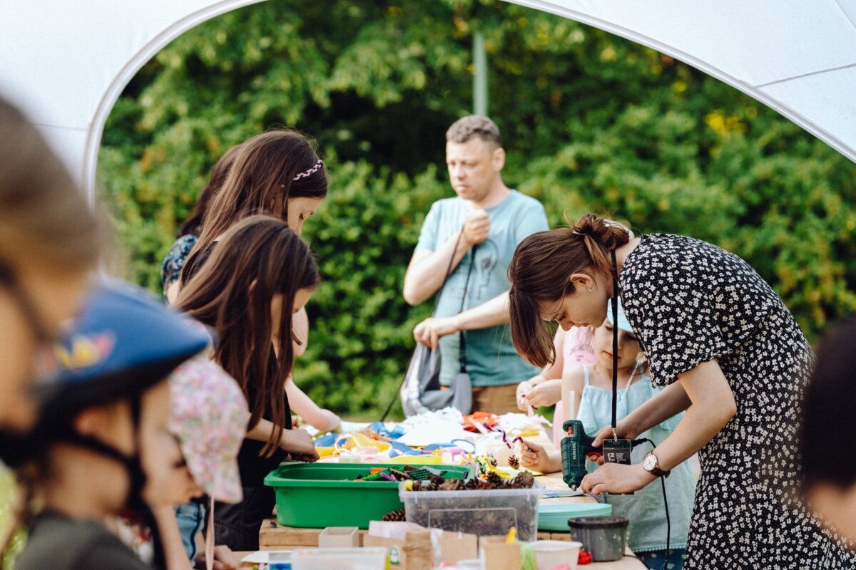 Children and adults take part in a handicraft activity during an outdoor event. A woman in a black dress with white floral prints helps a child with a handicraft project. Colorful materials and tools are spread out on tables. People focused on their activities under a white canopy, beautifully captured by a photographer from Warsaw.   