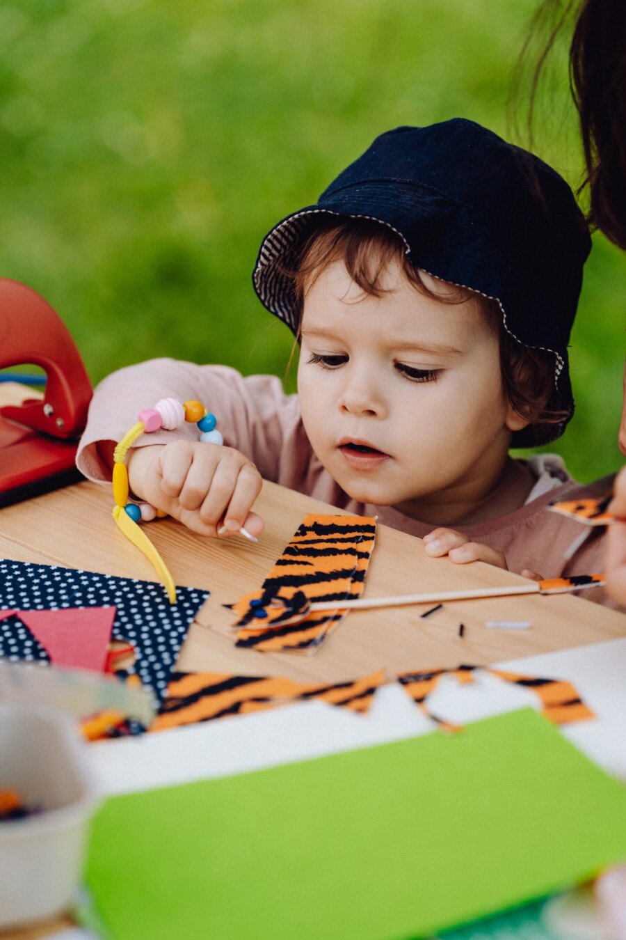 A young child in a blue hat and colorful beaded bracelet focuses on arts and crafts at an outdoor table. The table is set with vibrant paper and craft supplies. The blurry background, taken as part of the event photo shoot, suggests they are in a grassy park-like setting.  