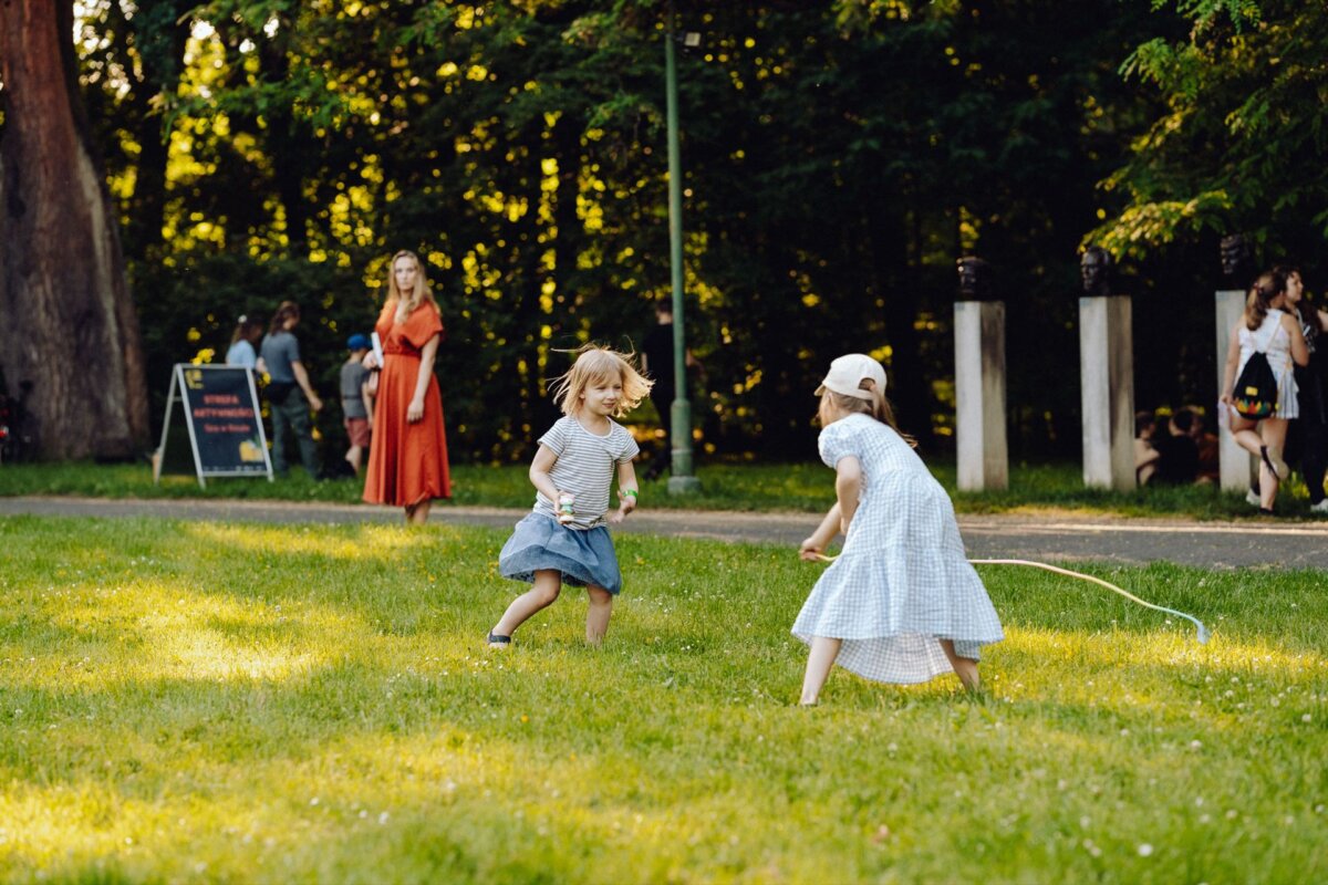 Two young girls are playing on a grassy field, one in a blue dress and the other in a white dress with a white cap, holding a skipping rope. In the background, captured by the photographer for the event, a woman in an orange dress is standing near a tree, and several people are scattered at the edge of the forest. 