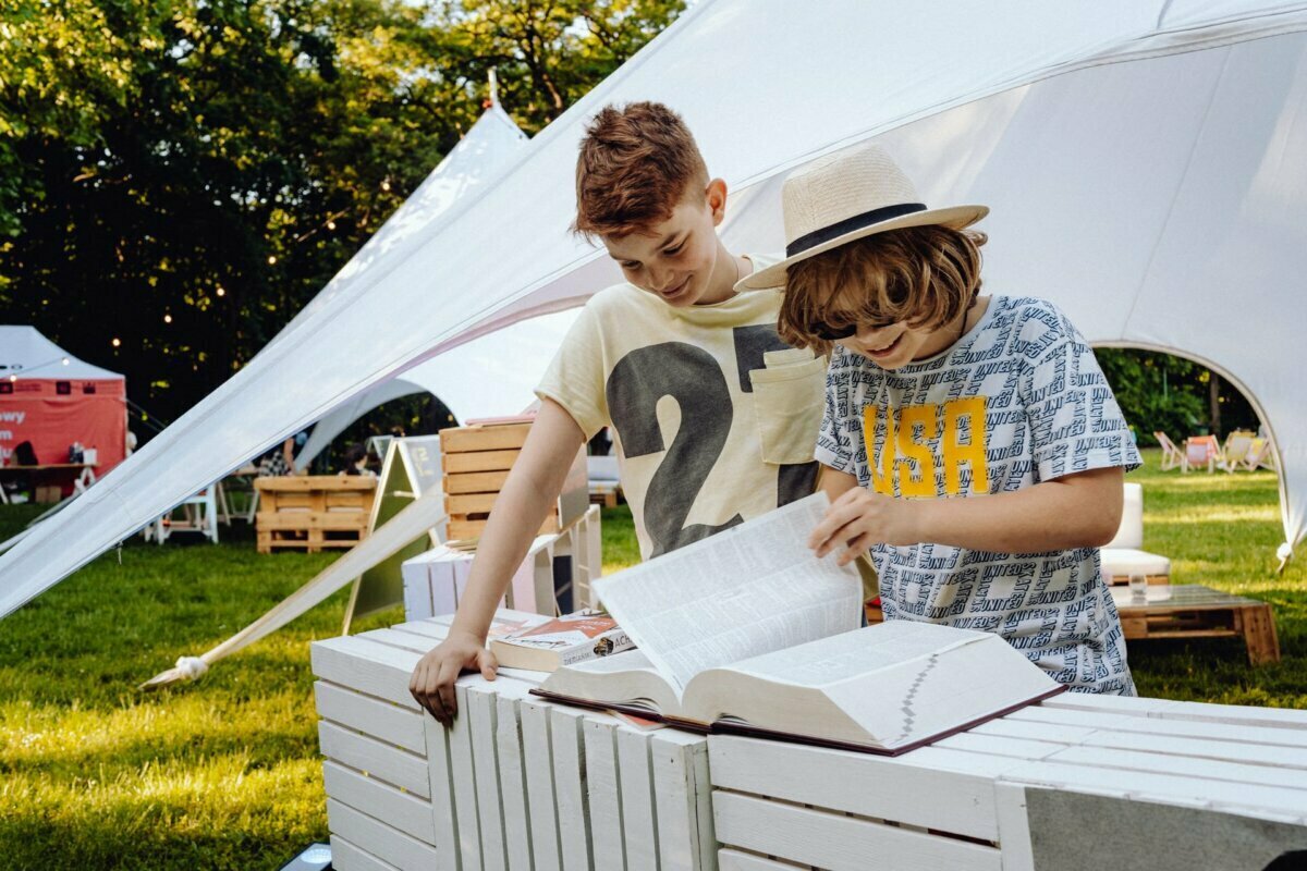 Two young boys are standing by an outdoor book display. One points to an open book, while the other looks at it. The scenery resembles a sunny park or festival, with modern tents and greenery in the background - a lovely scene captured by a photographer from Warsaw.  