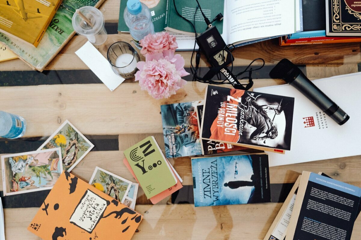 A cluttered table set with various books, postcards, a pink bouquet of flowers, a bottle of water, an electronic device and a microphone. The black-striped wooden surface alludes to the busy life of a photographer in Warsaw preparing to photograph an event. Some books are open, others are stacked or scattered.  