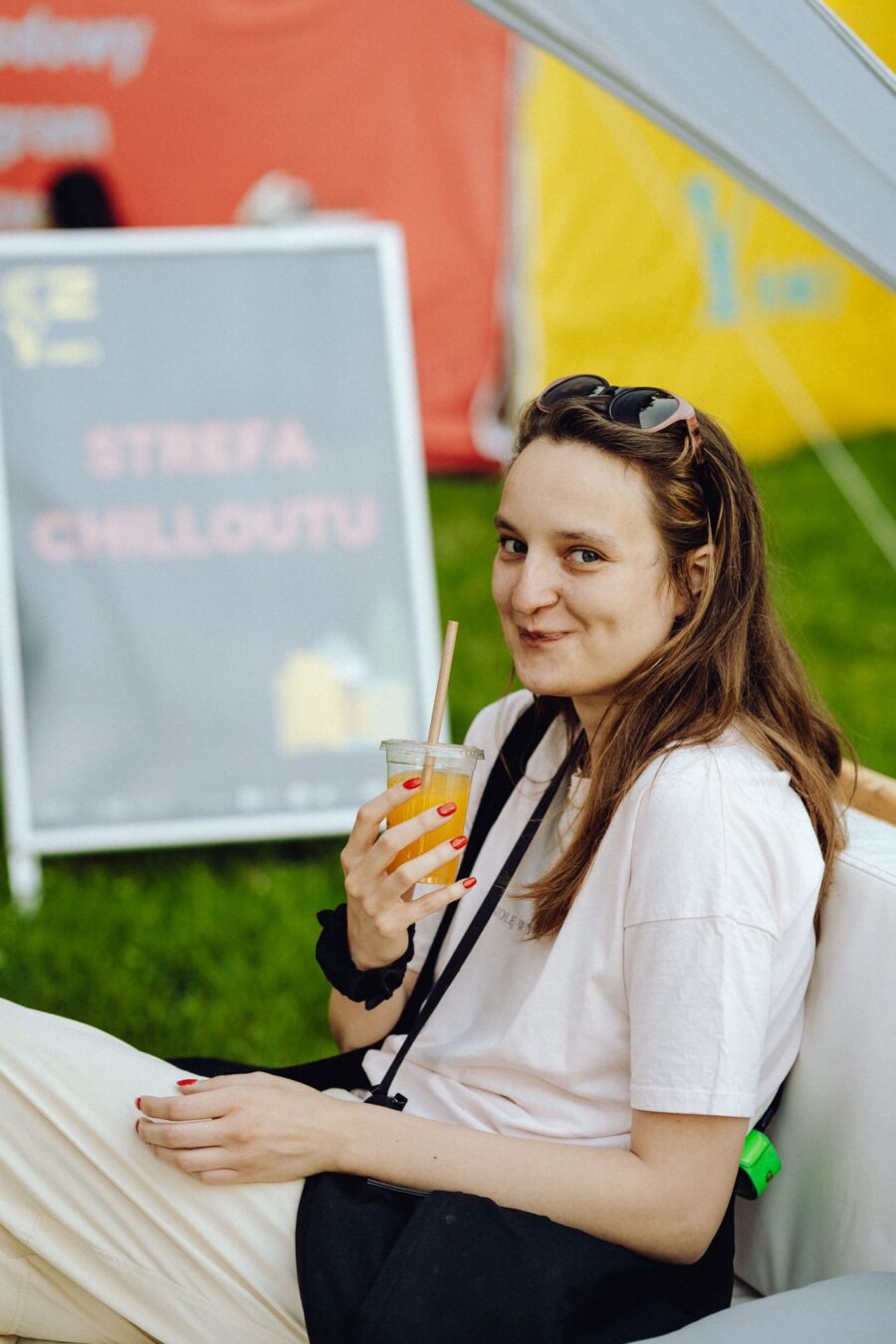 A young woman with long brown hair, sunglasses on her head and a bright T-shirt sits outside on a white couch. She smiles and holds a glass of orange juice through a straw. In the background you can see the sign "CHILLOUT ZONE," perfectly captured by event photographer Warsaw.  