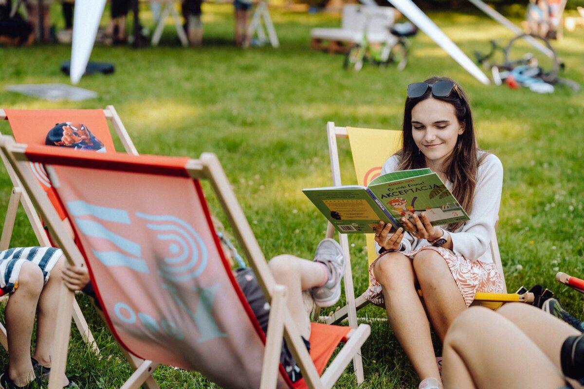 A young woman sits in an outdoor recliner and reads a children's book entitled "Acceptance" to the child next to her. Other children and adults enjoy the open grassy area in the background. The decorated deckchairs and relaxed atmosphere were beautifully captured by a photographer from Warsaw in this photo essay of the event.  