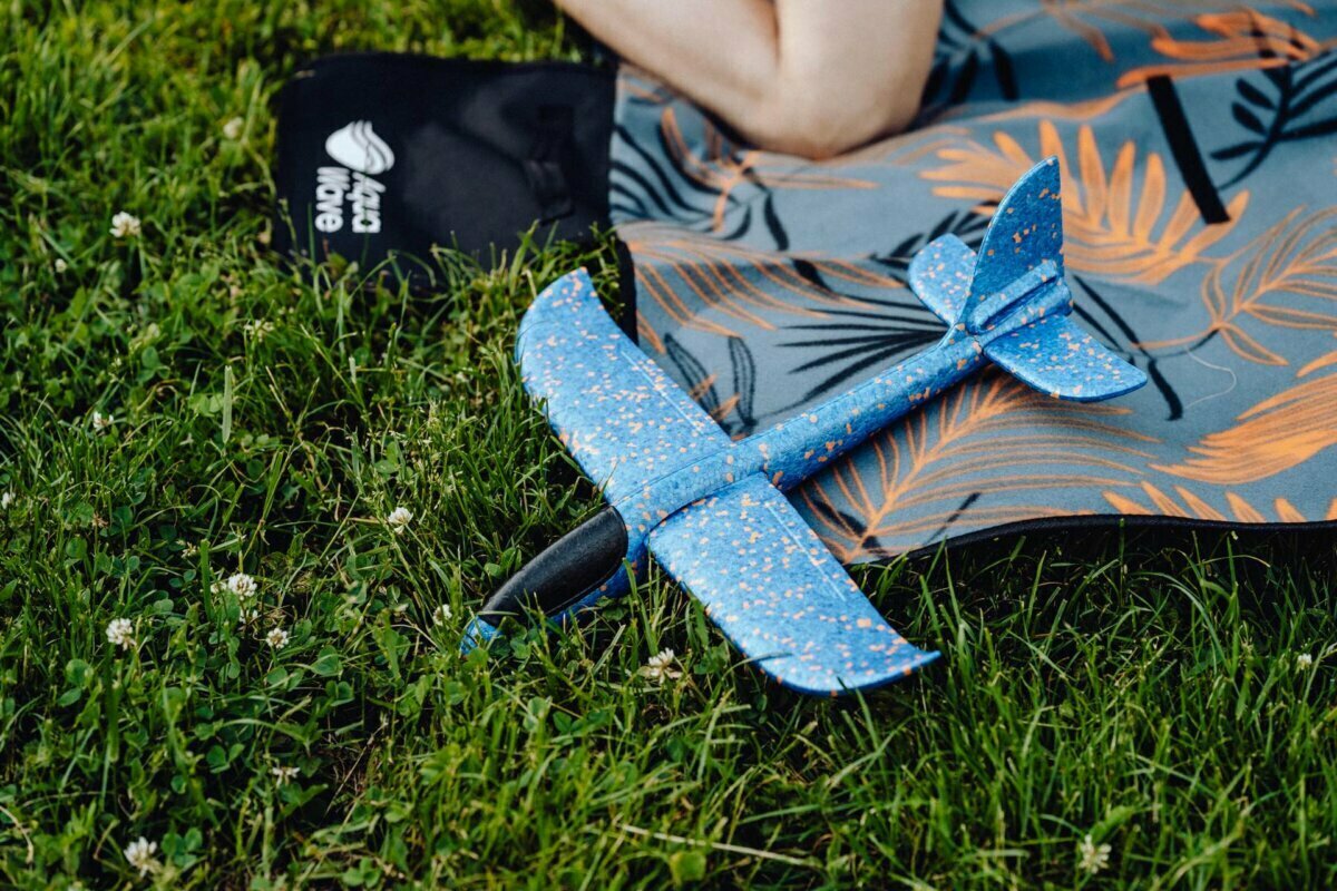 A blue spotted toy airplane with black accents sits on the grass next to a partially visible person wearing patterned clothing with leaves. A grassy area dotted with small white flowers enhances the scene, ideal for a photo-op of the event. 