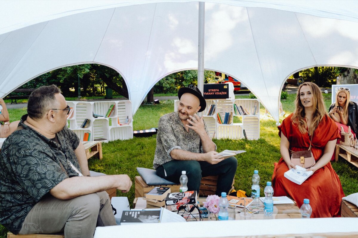 A group of three people are sitting under the white canopy of a tent at an outdoor event. They look busy in cheerful conversation. Around them are wooden boxes filled with books and a few bottles of water on a table. In the background are trees and greenery, perfectly captured by a photographer for an event from Warsaw.   