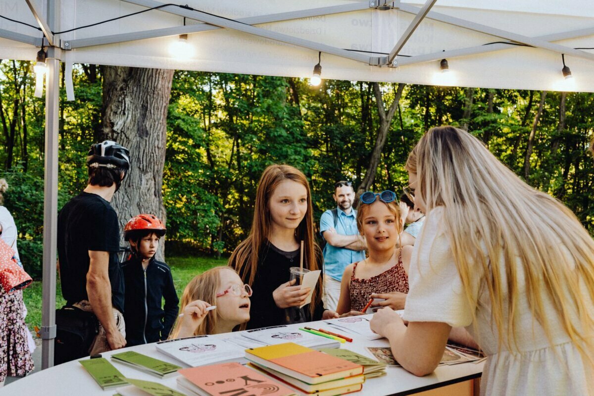 A group of two children and one adult are talking to a woman at an outdoor booth under a tent. Behind them, people are standing and talking against the backdrop of a lush green forest. The table is swamped with papers, notebooks and other items. The environment is bright and sunny, perfect for a photography event.   