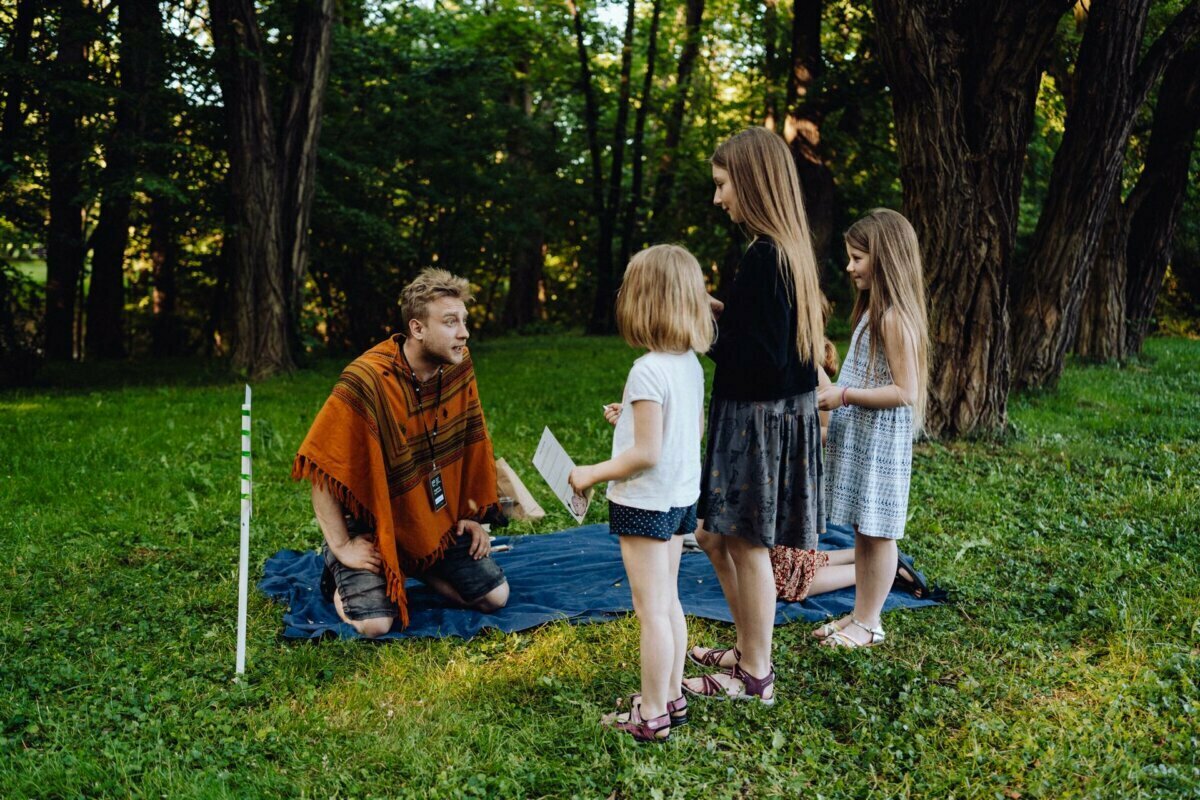 A person sitting on a blanket in a grassy, wooded area is holding a piece of paper and talking to three children standing around him. The children appear to be listening intently. Near the stage lies a walking stick, beautifully captured by event photographer Warsaw.  