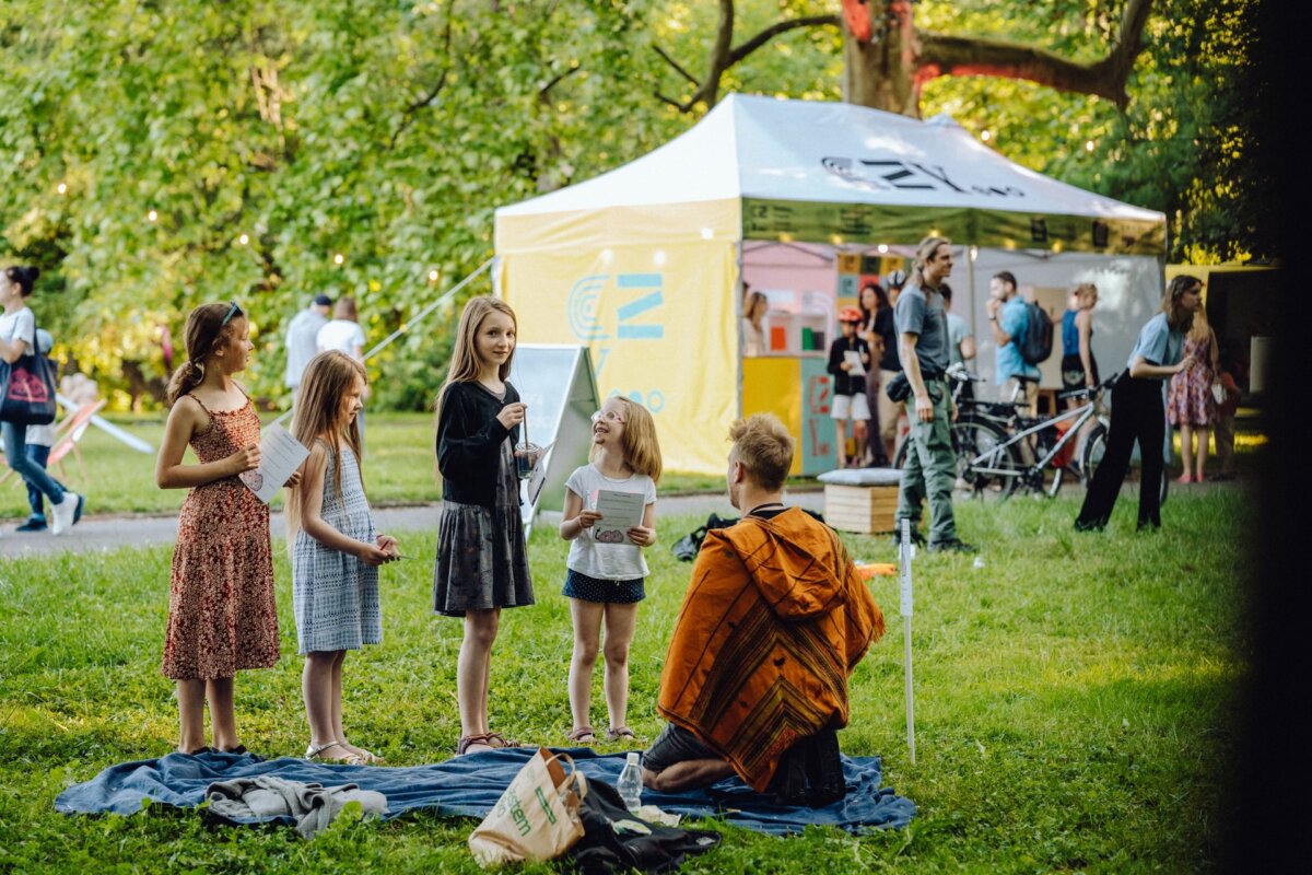 Four young girls gather on a blanket on the grass, reading or discussing something, while a person in an orange jacket kneels nearby. In the background is a yellow and white tent set up among the trees in the park, with other people loitering around - a perfect scene for a photo essay of the event. 