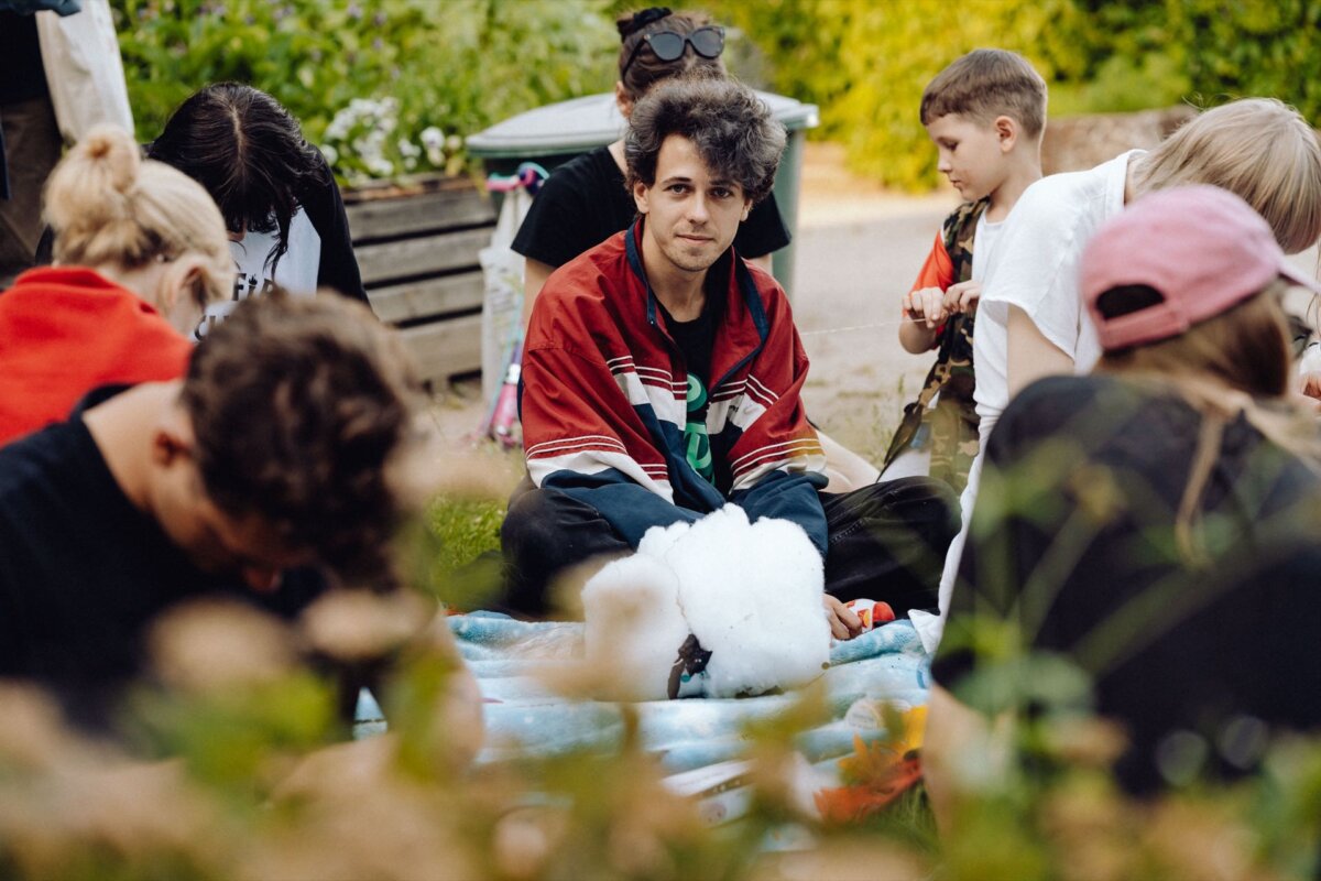 A group of people are sitting on the grass in the open air, busy with an activity or conversation. One person in the middle is looking into the camera, wearing a red and black jacket. Other people are focused on their tasks, against a backdrop of greenery and a wooden bench - a perfect photo-reportage of the event.  