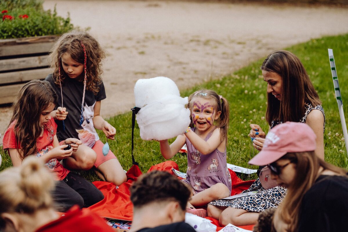 A group of children and an adult are sitting on a red blanket on the grass doing various activities. A girl with a painted face excitedly holds a large piece of cotton candy. The cheerful and lively scene perfectly captures the essence of the event photo report by a photographer from Warsaw.  