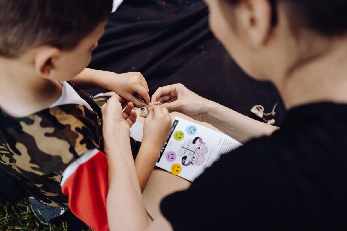 From the back, a child wearing a camouflage jacket and an adult can be seen working together on a small craft project. Both are focusing on string or thread, while the adult is holding an instruction manual with colorful smiley faces. This moment was perfectly captured by a photographer for an event from Warsaw.  