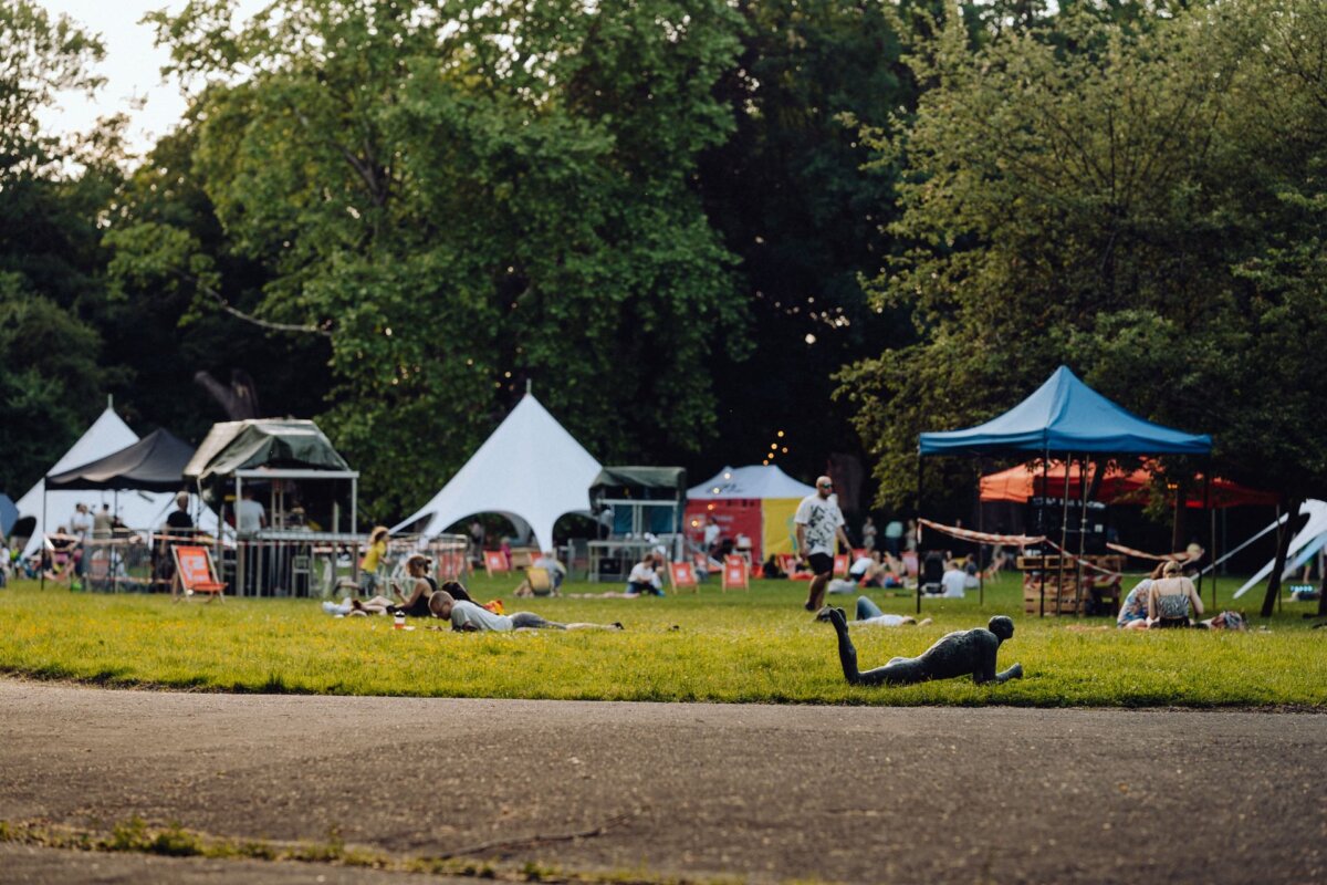 A casual party is taking place in a grassy park with a few white and blue tents. People are lounging on the grass and walking around, which was perfectly captured by a photographer from Warsaw. Trees in the background provide shade, and some signs and equipment are visible near the tents, adding to the relaxed atmosphere.  