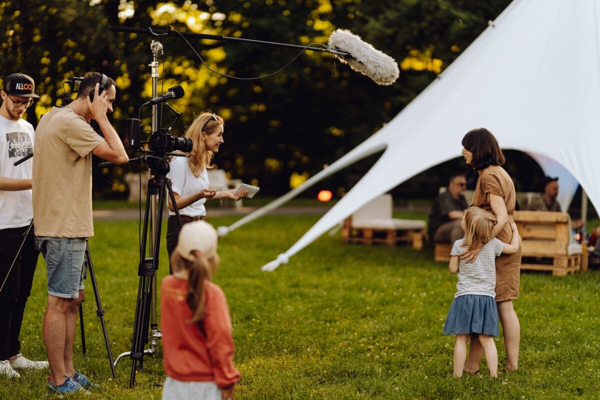 A film crew is shooting an outdoor scene with a woman and two children. The woman hugs the girl while the other child stands nearby. The crew uses a camera, boom microphone and other equipment. In the background, a white tent structure can be seen in a grassy field, perfect for photo coverage of the event.   