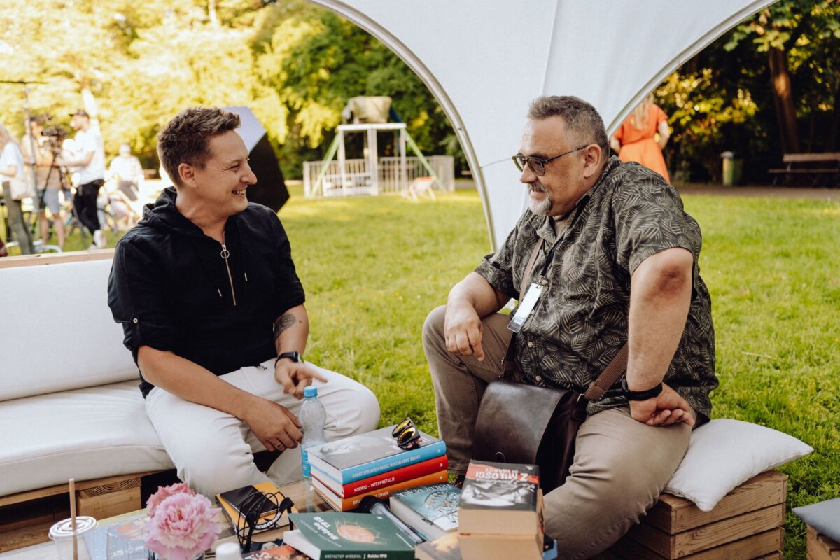 Two people are sitting on a white couch in the garden and having a conversation. The person on the left is smiling, wearing a black shirt. The person on the right is wearing a patterned shirt and glasses. In front of them on a table is a stack of books and flowers, captured by a photographer from Warsaw.   