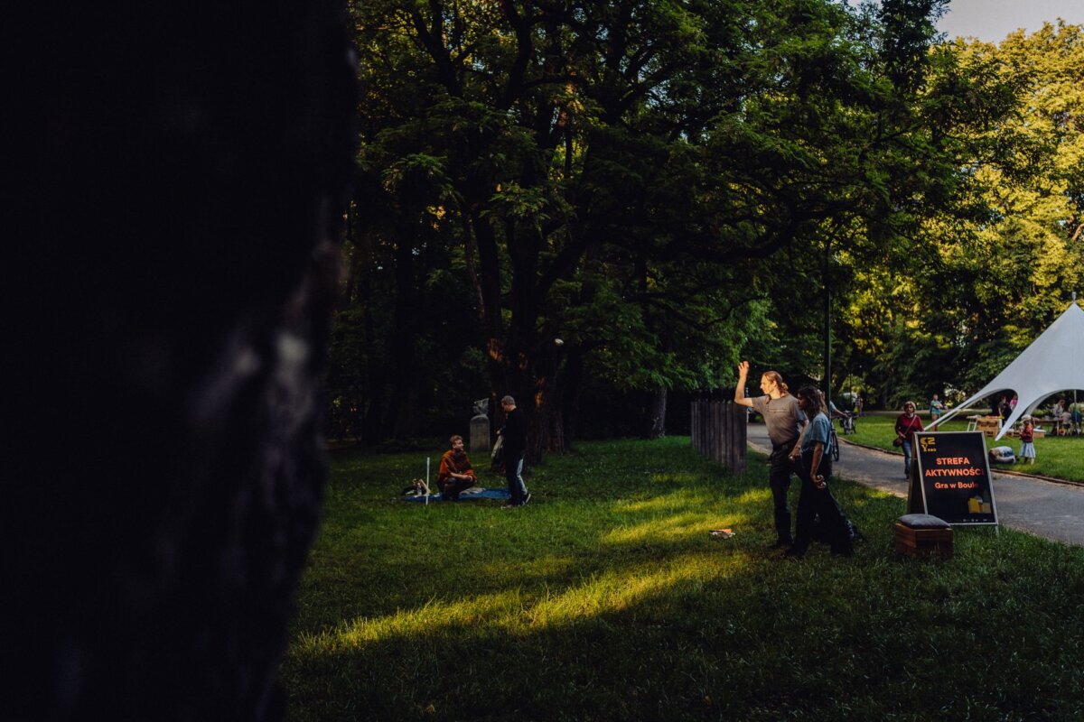 A man stands in a sunny park and gestures with his hand. Nearby, two people are sitting on the grass near the trees. In the background you can see a white tent and the sign "Activity Zone." The scene is calm, with patches of sunlight flying through the trees, which is perfect for capturing any photographic event in Warsaw.   