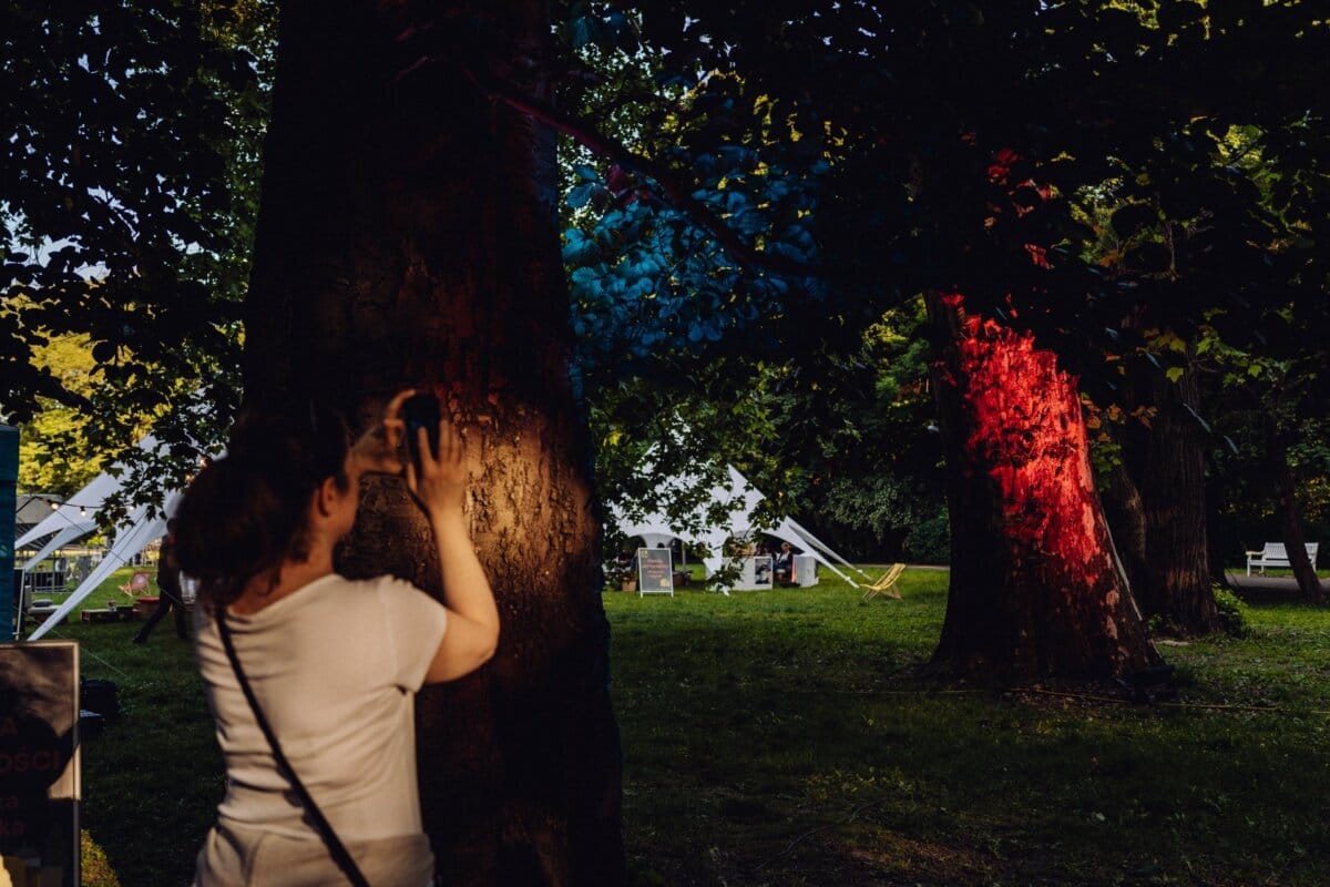 A woman in a white shirt and dark pants touches the trunk of a large tree in a park at dusk. The tree is illuminated with red and blue lights, creating a wonderful backdrop for *photographs for an event*. Tents and trees can be seen in the background, and the ground is overgrown with grass.  