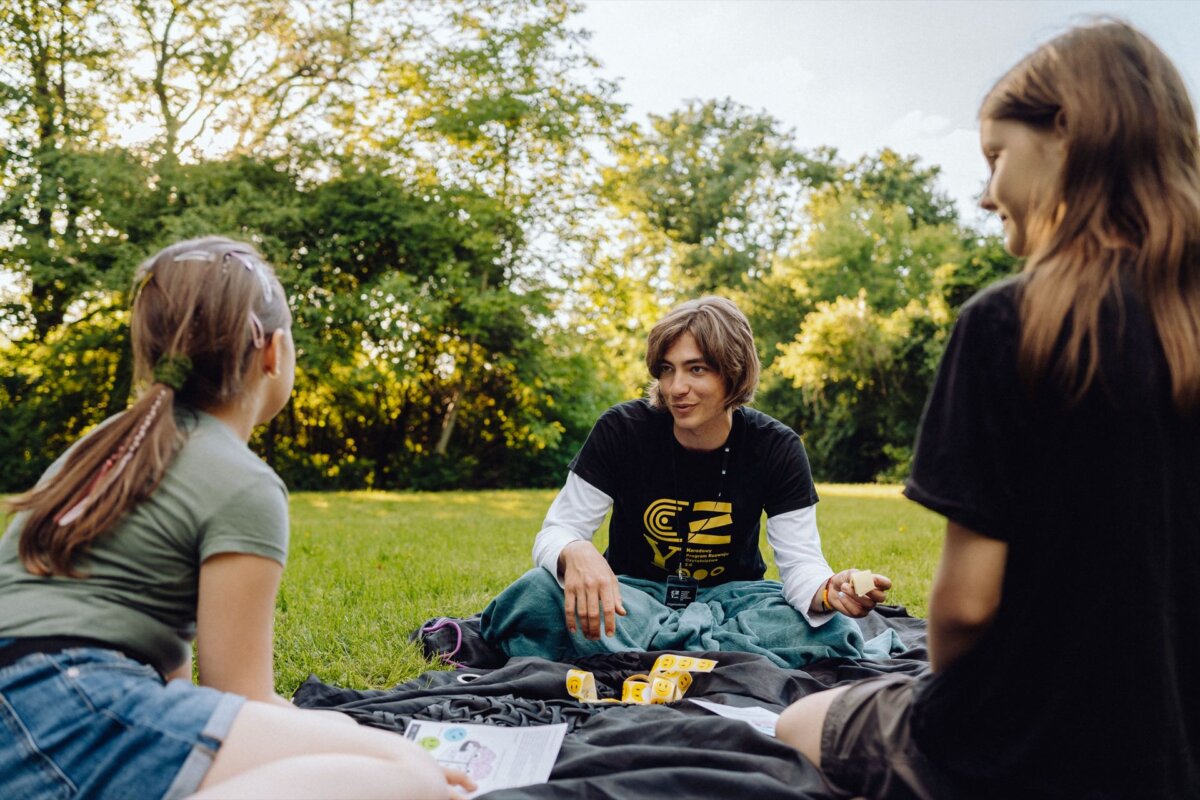 Three people are sitting on a blanket in the park, talking and playing dice. They are surrounded by lush greenery and the sun is shining brightly. One gets the impression that this is a scene captured by an event photographer Warsaw, where one person holding a dice is talking to the other two, who are listening intently.  