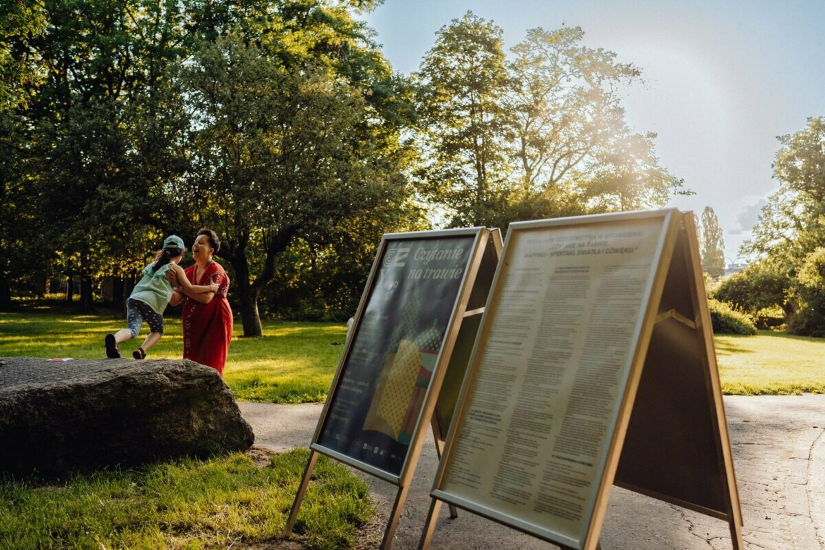 A woman in a red dress and a child play and laugh near a large rock in a sunny park with lush green trees. In the foreground are two large signs displaying information. Sunlight filters through the trees, casting a warm glow on the grassy area - perfect for a photo essay of the event.  