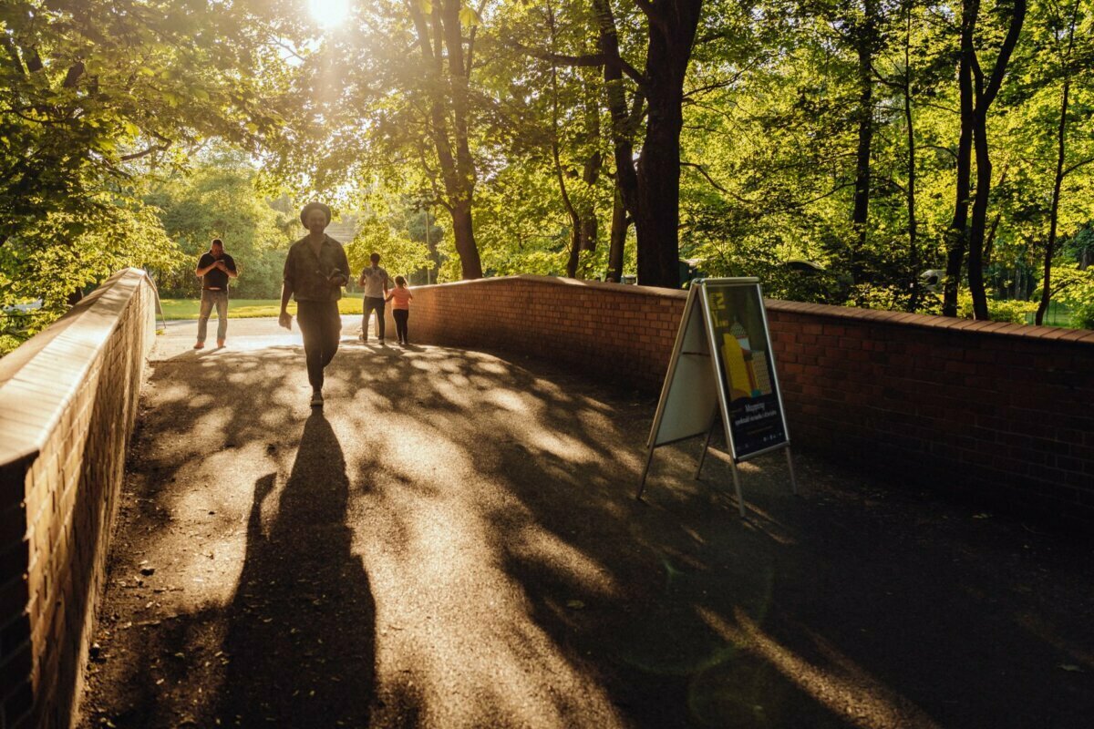 A person walks along a brick path through a sun-soaked park. Sunlight pierces through the treetops, casting long shadows on the ground. Two other people are walking in the background. A signboard placed on the right side of the path captures the moment like a photo report from an event by an event photographer Warsaw.   