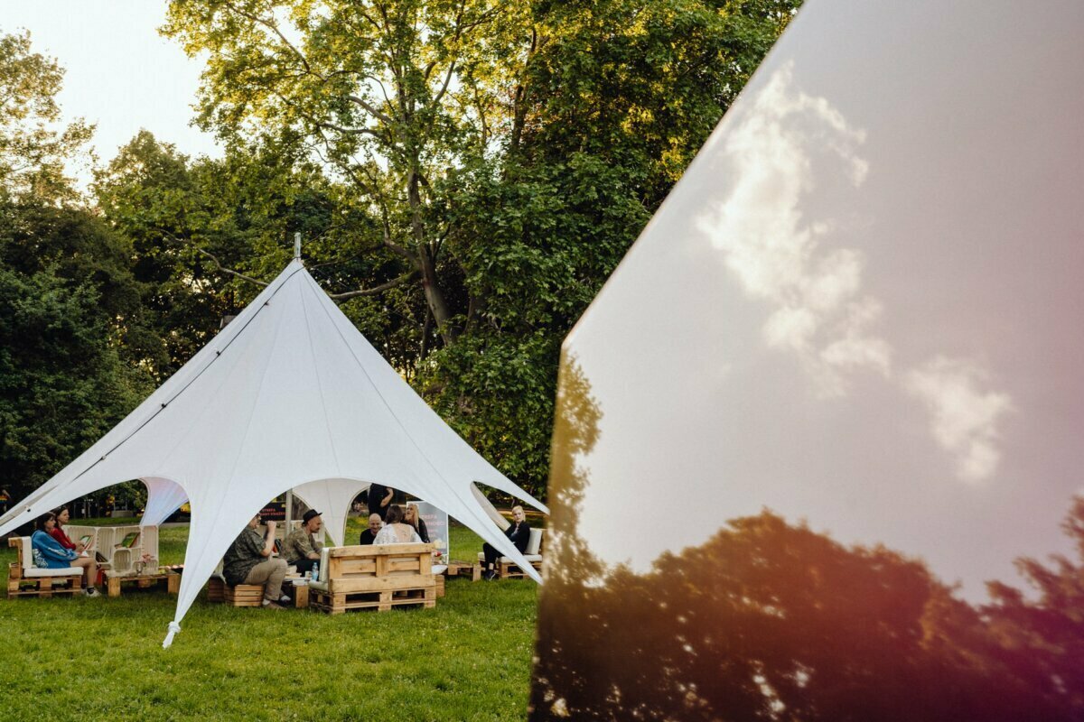 A group of people gather under a large white canopy tent set up in a grassy, tree-lined park. Wooden benches and tables made of pallets are set up under the tent. Sunlight filters through the trees, creating a peaceful, natural setting - perfect for an event photo essay by a talented event photographer from Warsaw.  