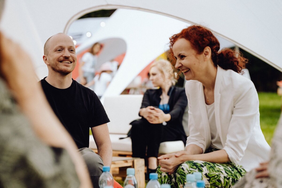 A group of people gathered outside under a white tent. In the foreground is a smiling and chatting man in a black shirt and a red-haired woman in a white jacket, beautifully captured by a photographer from Warsaw. In the background, a woman with blond curly hair sits on a white couch and looks away.  