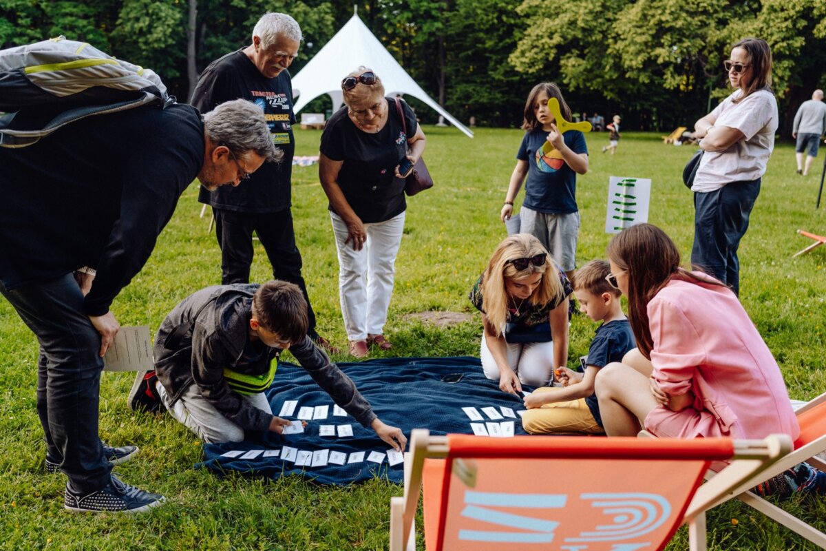 A group of people gather outdoors on a grassy area, engaging in an activity of spreading cards on a dark blanket. They appear to be in a park with trees and a large white tent in the background. Some are standing, others are sitting or kneeling. It's ideal for photo coverage of the event.   