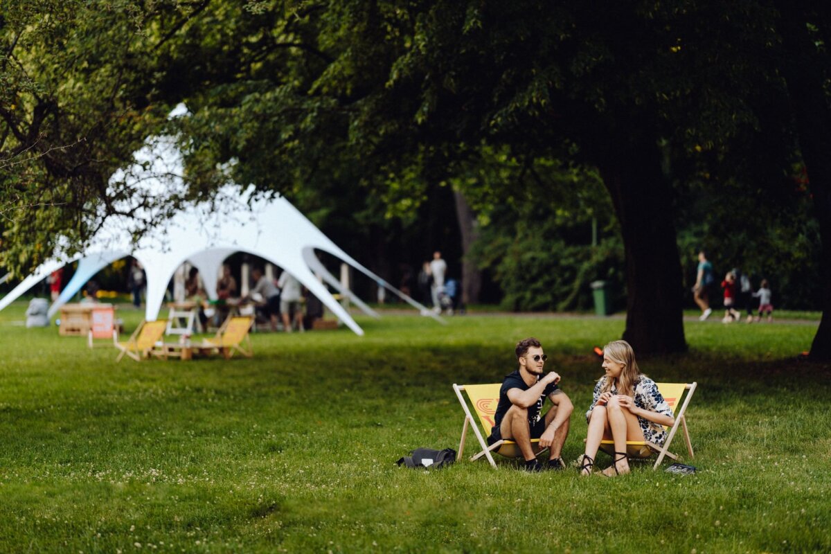 A man and a woman are sitting on deck chairs and talking in a park. A white tent is set up behind them, and additional people can be seen in the background enjoying the greenery and outdoor environment. The scene is reminiscent of a perfect photo-op of an event on a sunny day with lush green trees and grass.  