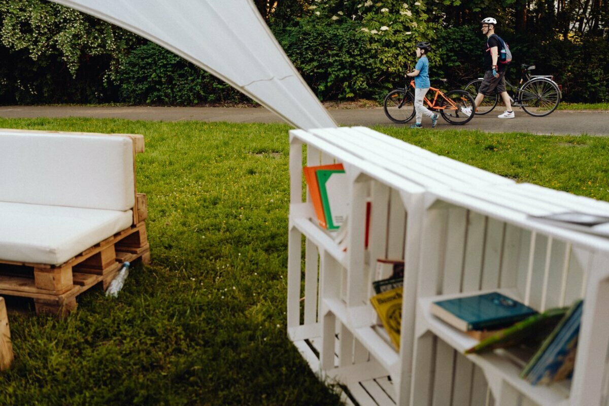 The cozy outdoor space includes a white couch and a bookshelf with colorful books. Outside the setting, a man and a child are walking their bicycles along a path in the park, surrounded by lush greenery. This peaceful moment, skillfully captured by the photographer for the event, is partially obscured by the white canopy on the left.  