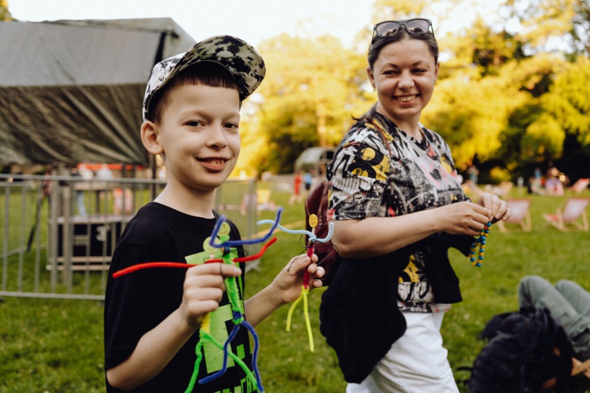 A smiling boy in a camouflage cap and black t-shirt presents a colorful pipe-cleaning artwork, while the woman standing next to him, also smiling, holds a bead craft. They are outdoors, with green grass and trees in the background, enjoying the sunny day - perfect for a photo essay of the event. 