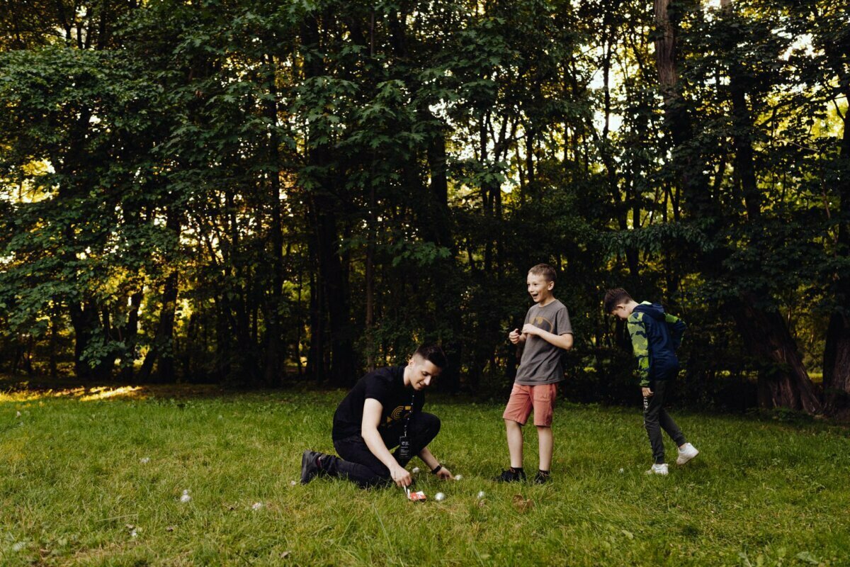 A person kneels on the grass in a wooded area, showing something to two children standing nearby. The perfect setting for an event photographer Warsaw is a sunny forest with lush greenery in the background. The two children are watching intently and all three seem to be engaged in the activity.  