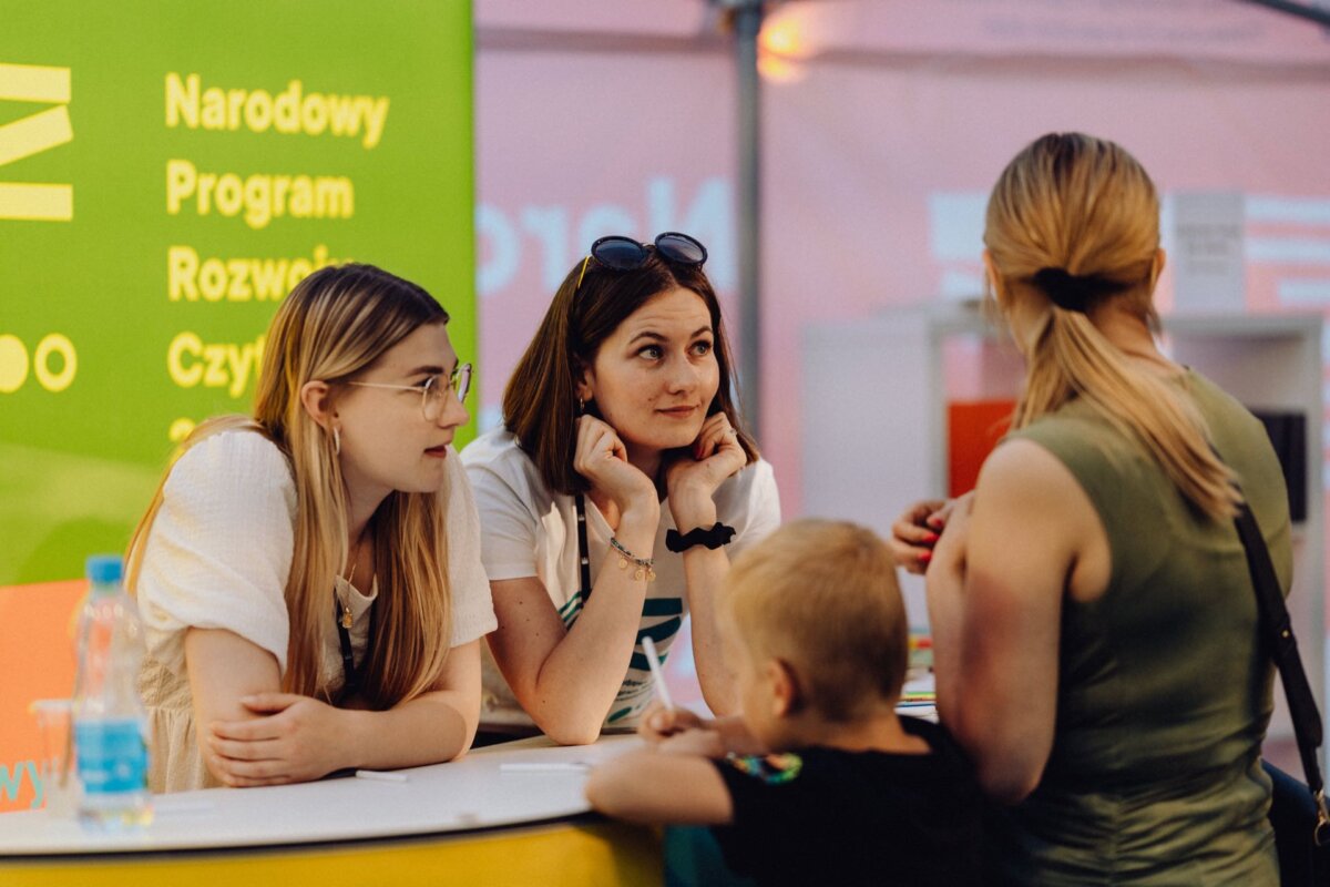 There are two women at the booth, potentially giving information to a woman with a child standing on the other side of the counter. The booth is decorated with a green and yellow background with the Polish text "National Reading Development Program." This photo report of the event captures the vibrant atmosphere of the fair.  