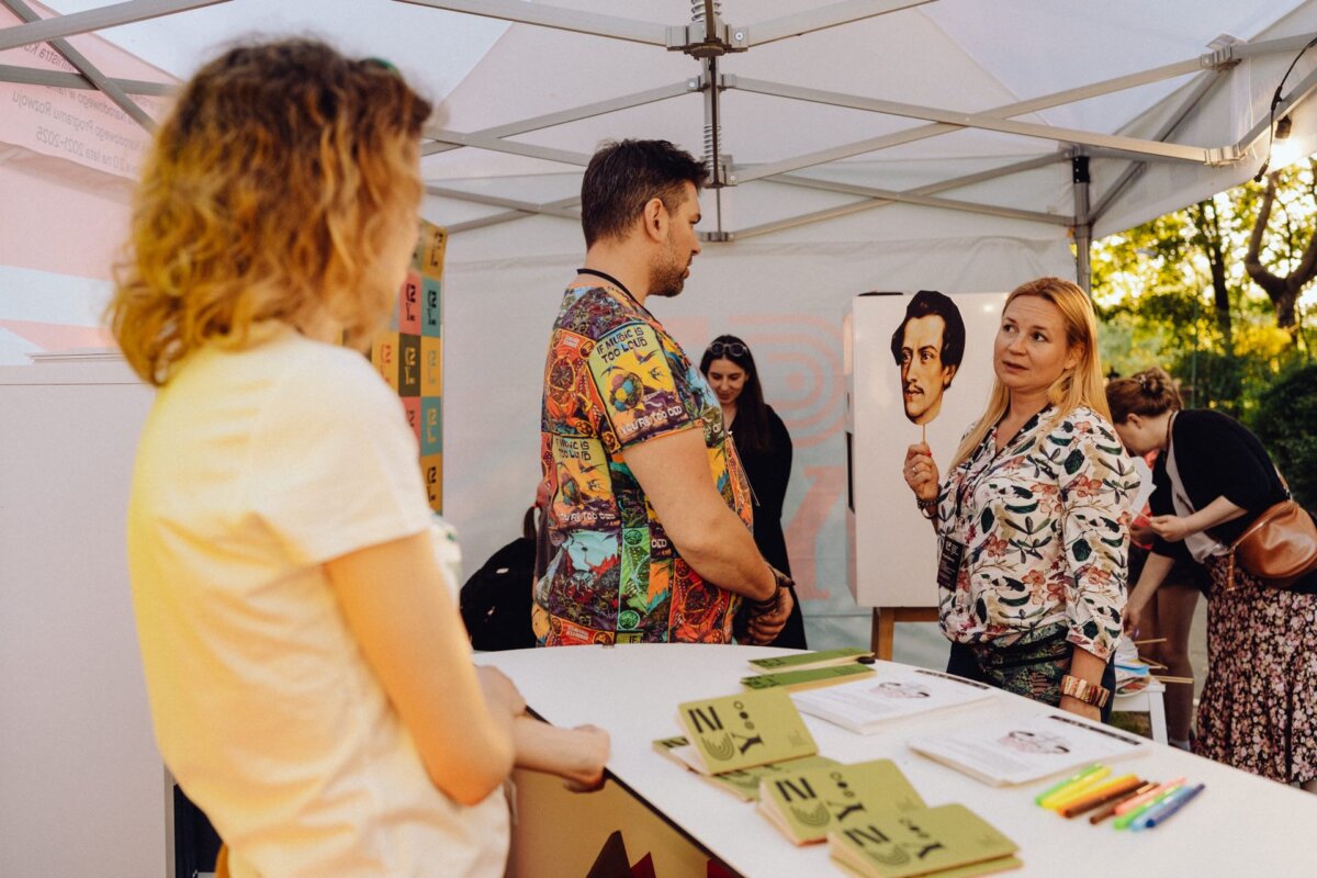 A woman stands under a tent, holding a sketch of a man, and talks to a man in a collared shirt. Another woman with curly hair watches from behind a table with various notepads and pens, capturing the moment for a *photo report of the event*. More people and artwork can be seen in the background.  