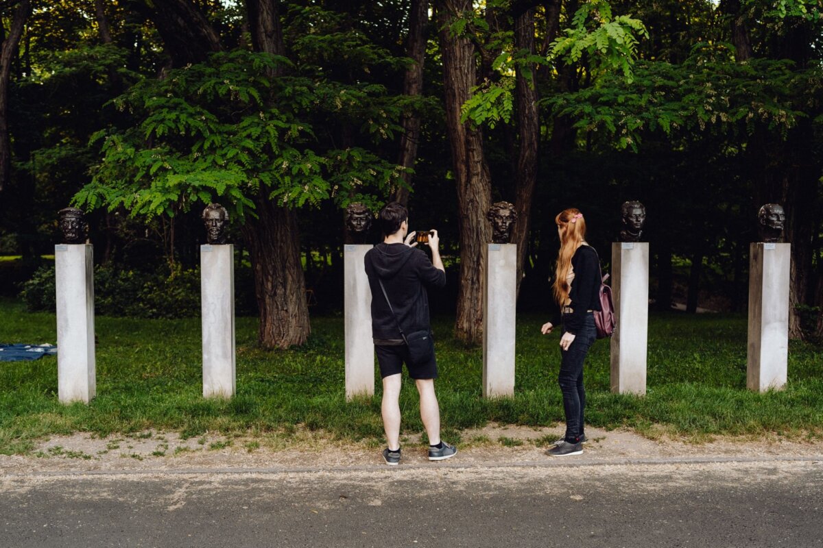 Two people stand in front of an exhibition of several sculptures of heads mounted on white pedestals, outdoors, surrounded by trees. A photographer at the event takes pictures of the sculptures, while the other looks at them. 