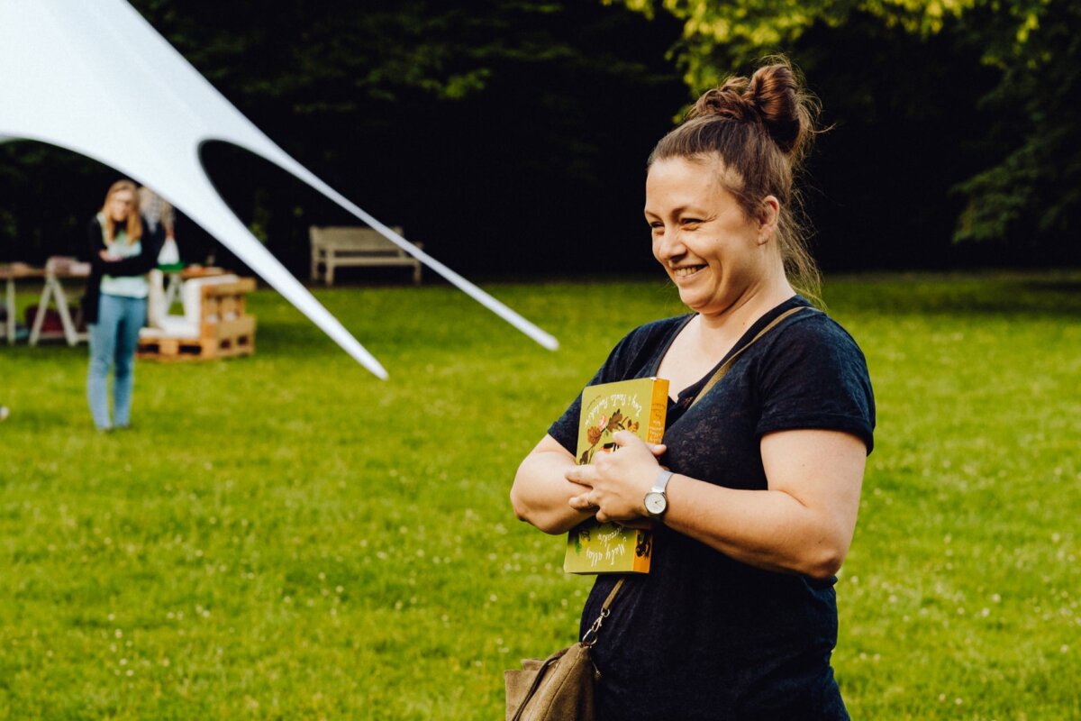 A smiling woman with brown hair tied in a top knot holds two books while standing on a grassy area at an outdoor event. She is wearing a black shirt and a watch. In the background is a white canopy tent, a bench, and another person in the distance. This photo shows a snapshot of the event by a photographer from Warsaw.   