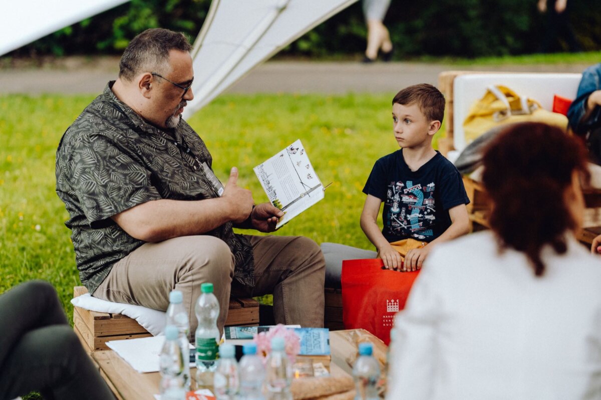 A man is reading a book, showing the pages to a young boy. They are sitting outside on wooden benches, surrounded by several bottles and objects on a low table. Other people are partially visible, indicating a casual outdoor gathering - perfect for a photo essay of the event by a skilled event photographer.  